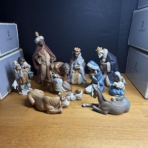 May include: A collection of porcelain nativity figurines, including the three wise men, Mary, Joseph, and baby Jesus, along with animals. The figures are in various colors and poses, set on a wooden surface. Boxes are stacked in the background.