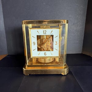 May include: A gold-toned rectangular clock with a white face and black numerals. The clock's face displays the numbers 12, 3, 6, and 9. The clock is encased in glass and has a visible pendulum.