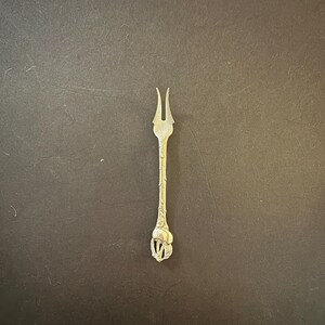 May include: A silver-coloured, two-pronged fork with a decorative handle. The handle has an ornate design, possibly floral or organic, and the fork is set against a dark background.