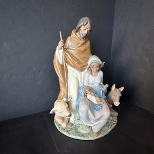 Può includere: Un set di figurine della natività in porcellana che rappresenta Giuseppe, Maria e Gesù bambino. Le figurine sono vestite con abiti tradizionali e sono circondate da un asino e una mucca. La scena è ambientata su una base verde.