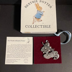 Peut inclure: Une décoration en argent représentant un personnage de la série de livres "Le Monde de Beatrix Potter" de Beatrix Potter. La décoration représente une grenouille portant un chapeau rouge et tenant une canne à pêche, avec un poisson au bout de la ligne. La décoration est sur un fond de velours rouge. Le texte "The World of Beatrix Potter" est visible sur la carte en arrière-plan.
