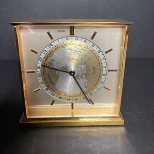 Pode incluir: Um relógio de mesa dourado com um mostrador de mapa-múndi. O relógio tem uma agulha das horas e dos minutos preta e uma agulha dos segundos branca. O mostrador do relógio também tem os nomes de cidades de todo o mundo, incluindo Paris, Cairo, Moscou e Bombaim.