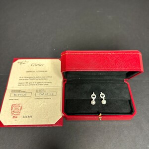 May include: A red Cartier box with a white pearl and diamond earring set inside. The box is open and the earrings are visible. The certificate for the earrings is visible next to the box. The certificate is white with black text and reads 'Cartier CERTIFICAT/CERTIFICATE En or 18 carats ou en platine, nous certifions que ce bijou est authentique. Cartier jewel Made in 18K gold or in platinum, we certify that this is an authentic Cartier jewel. 518915 Feb 13-07 Cartier 585898'