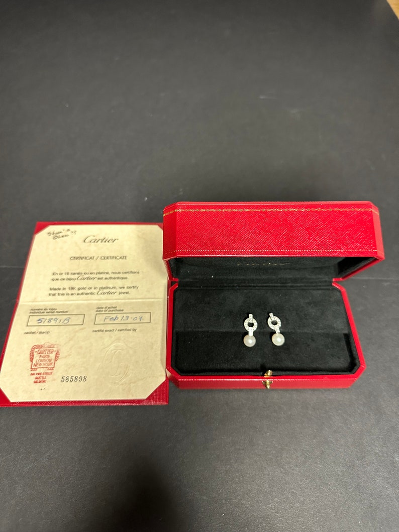 May include: A red Cartier box with a white pearl and diamond earring set inside. The box is open and the earrings are visible. The certificate for the earrings is visible next to the box. The certificate is white with black text and reads 'Cartier CERTIFICAT/CERTIFICATE En or 18 carats ou en platine, nous certifions que ce bijou est authentique. Cartier jewel Made in 18K gold or in platinum, we certify that this is an authentic Cartier jewel. 518915 Feb 13-07 Cartier 585898'