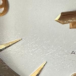 Puede incluir: Primer plano de una esfera de reloj blanca con marcadores de hora y manecillas de color dorado. La esfera del reloj tiene una superficie texturizada y el n&uacute;mero 9 es visible.