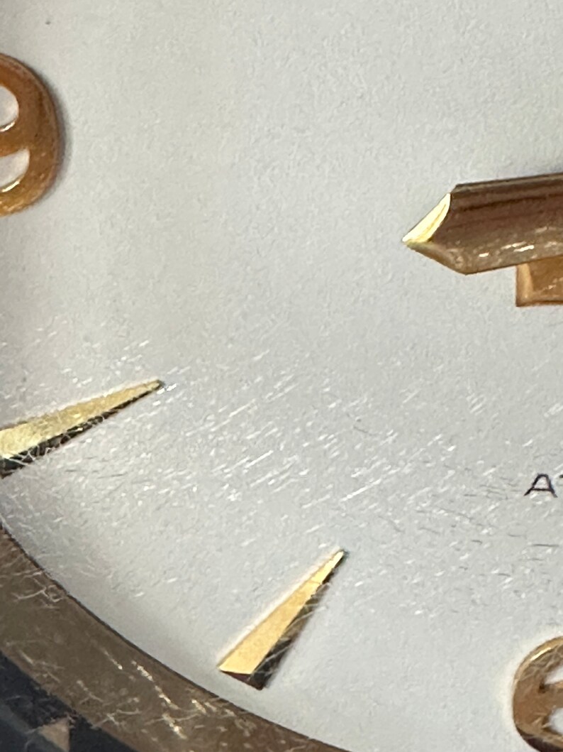 Puede incluir: Primer plano de una esfera de reloj blanca con marcadores de hora y manecillas de color dorado. La esfera del reloj tiene una superficie texturizada y el n&uacute;mero 9 es visible.