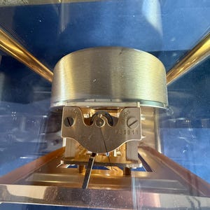May include: Close-up of a brass-colored Atmos clock mechanism. The clock features a gold-colored frame and a clear glass enclosure. The mechanism includes a brass-colored balance wheel and a small plate with the number 48811.