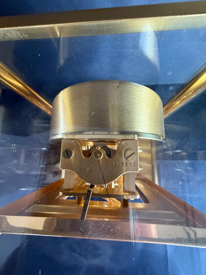 May include: Close-up of a brass-colored Atmos clock mechanism. The clock features a gold-colored frame and a clear glass enclosure. The mechanism includes a brass-colored balance wheel and a small plate with the number 48811.