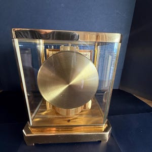 May include: A gold-toned, rectangular clock with a glass enclosure. The clock features a large, circular pendulum and a gold-colored base and frame. The clock's face is visible through the glass, showcasing its intricate design.