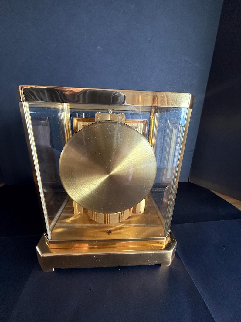 May include: A gold-toned, rectangular clock with a glass enclosure. The clock features a large, circular pendulum and a gold-colored base and frame. The clock's face is visible through the glass, showcasing its intricate design.