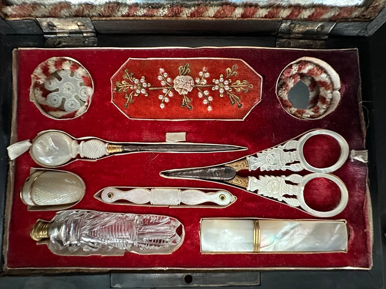 Pu&ograve; includere: Un kit da cucito vintage con un interno in velluto rosso. Il kit comprende un paio di forbici, un ago, un ditale, una piccola bottiglia e una piccola custodia. Il kit &egrave; decorato con un motivo floreale.