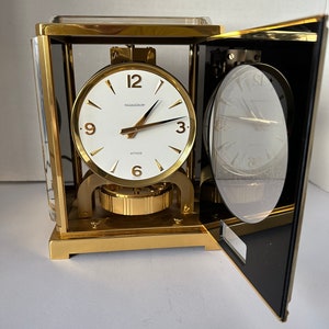 Puede incluir: Un reloj anal&oacute;gico rectangular de color dorado con una esfera blanca y agujas negras. El reloj est&aacute; etiquetado como "Atmos" y "Jaeger-LeCoultre". El reloj est&aacute; abierto, revelando el mecanismo interno.