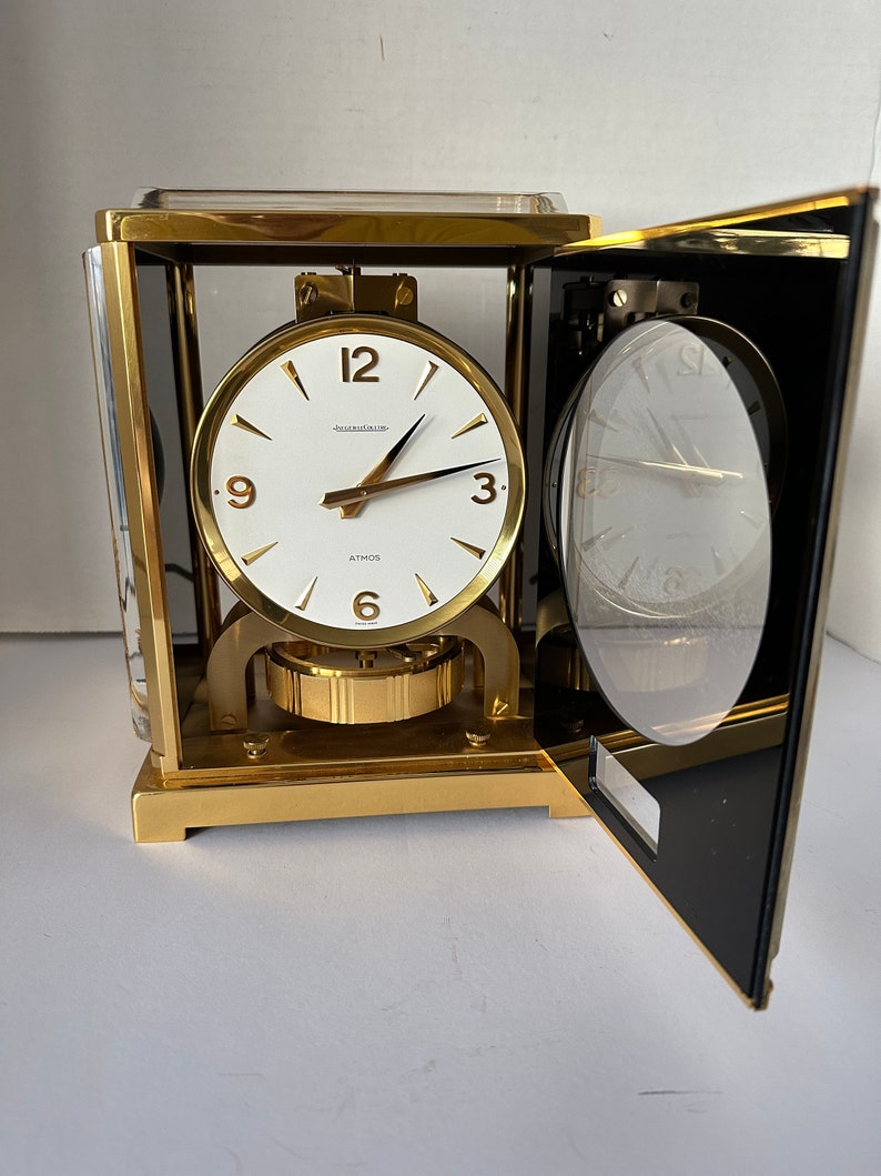 Puede incluir: Un reloj anal&oacute;gico rectangular de color dorado con una esfera blanca y agujas negras. El reloj est&aacute; etiquetado como "Atmos" y "Jaeger-LeCoultre". El reloj est&aacute; abierto, revelando el mecanismo interno.