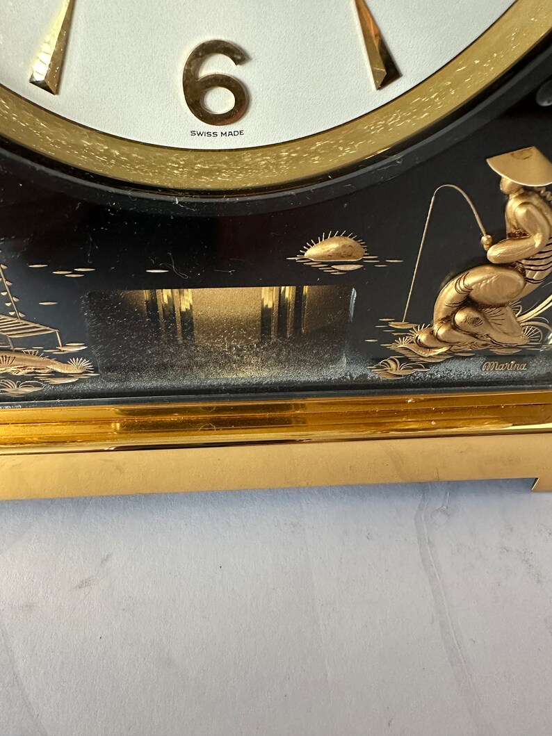 Puede incluir: Un reloj negro y dorado con un borde dorado y un dise&ntilde;o de pescador. La esfera del reloj es blanca con n&uacute;meros romanos negros y las palabras "Swiss Made".