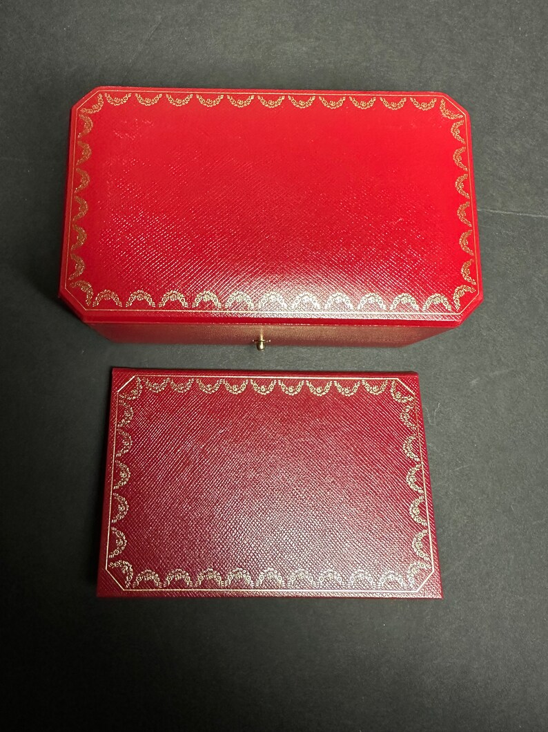 May include: Two red leather boxes with gold trim. The top box is slightly larger than the bottom box. Both boxes have a repeating pattern of gold crescents around the edges.