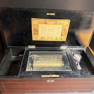 May include: An antique music box with a dark wood exterior and a hinged lid. The interior features a glass-covered mechanism and a decorative label with handwritten text. The box is open, revealing the inner workings.