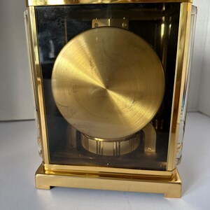 Puede incluir: Un reloj de color dorado con una caja de cristal y un mecanismo de relojer&iacute;a visible. El reloj tiene una forma cuadrada y un acabado dorado pulido.
