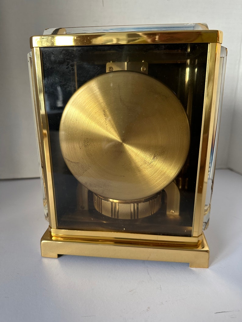 Puede incluir: Un reloj de color dorado con una caja de cristal y un mecanismo de relojer&iacute;a visible. El reloj tiene una forma cuadrada y un acabado dorado pulido.