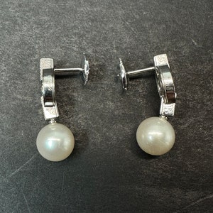May include: A pair of silver earrings with a pearl drop and a small diamond-like design on the top. The earrings are on a gray surface.