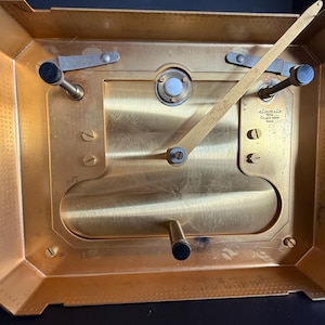 May include: A close-up view of the back of a gold-toned clock mechanism. The clock has a rectangular shape with a series of gears, levers, and screws. The clock's face is not visible. The text "J.C. Collet & Co. Calibre Luxe Swiss" is visible.