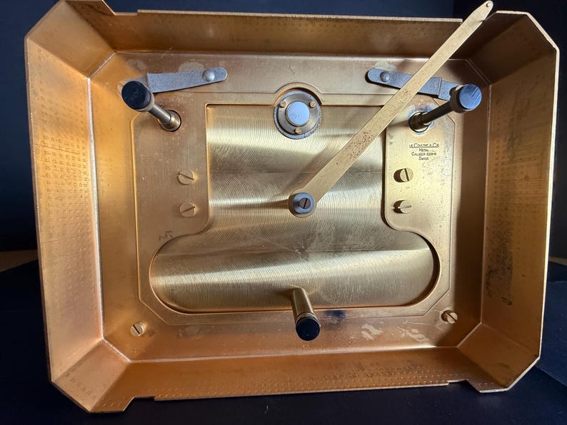 May include: A close-up view of the back of a gold-toned clock mechanism. The clock has a rectangular shape with a series of gears, levers, and screws. The clock's face is not visible. The text "J.C. Collet & Co. Calibre Luxe Swiss" is visible.