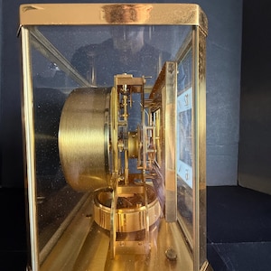 May include: A gold-toned, rectangular clock with a glass enclosure. The clock's internal mechanism is visible, featuring gears and a pendulum. The clock face is white with black numerals. The clock sits on a gold base.