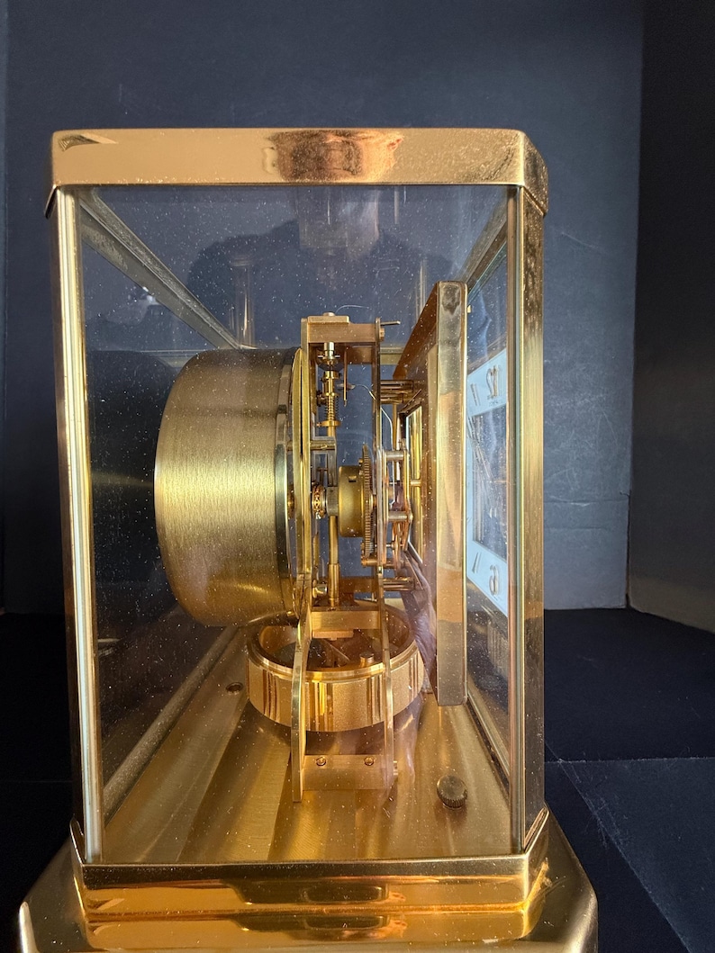 May include: A gold-toned, rectangular clock with a glass enclosure. The clock's internal mechanism is visible, featuring gears and a pendulum. The clock face is white with black numerals. The clock sits on a gold base.