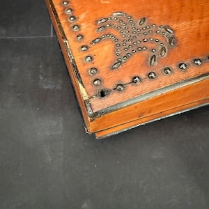 Pu&ograve; includere: Una scatola di legno con un design decorativo di borchie metalliche a forma di palme. La scatola ha una finitura marrone e una maniglia in metallo.