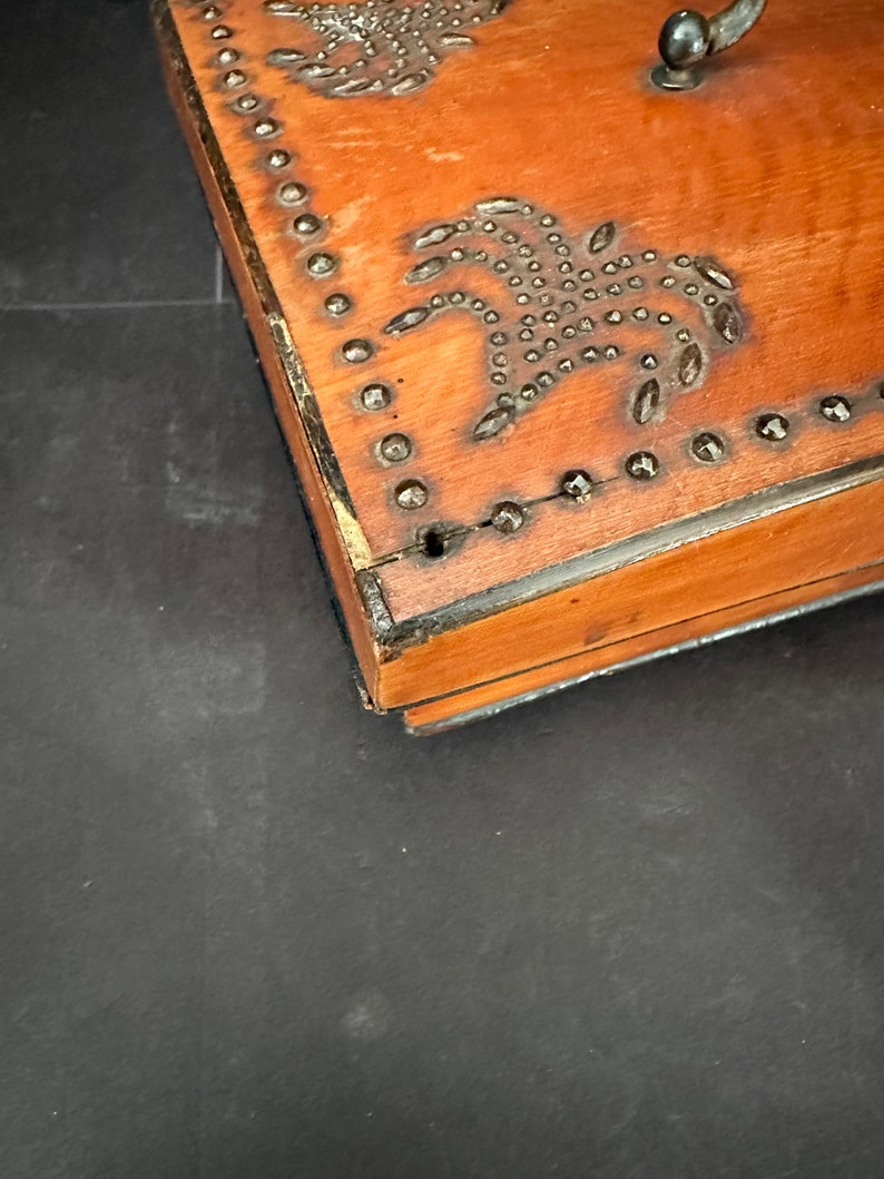 Pu&ograve; includere: Una scatola di legno con un design decorativo di borchie metalliche a forma di palme. La scatola ha una finitura marrone e una maniglia in metallo.