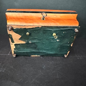 Pu&ograve; includere: Una scatola di legno con un rivestimento in feltro verde scuro e un buco della serratura nella parte superiore. La scatola ha quattro piedini in ottone e una chiave in ottone nel buco della serratura.