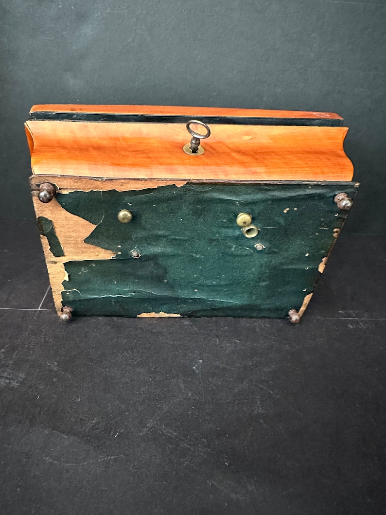 Pu&ograve; includere: Una scatola di legno con un rivestimento in feltro verde scuro e un buco della serratura nella parte superiore. La scatola ha quattro piedini in ottone e una chiave in ottone nel buco della serratura.