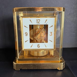 May include: A gold-toned Atmos clock with a white square clock face. The clock face has black numerals and hands. The clock is encased in a clear glass box. The clock's mechanism is visible. The clock sits on a gold base.