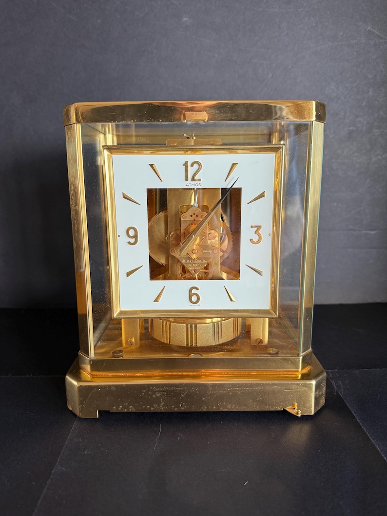 May include: A gold-toned Atmos clock with a white square clock face. The clock face has black numerals and hands. The clock is encased in a clear glass box. The clock's mechanism is visible. The clock sits on a gold base.