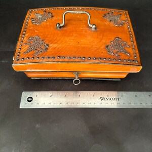 Pu&ograve; includere: Una scatola di legno con un coperchio incernierato, una maniglia in metallo e un buco della serratura in metallo. La scatola &egrave; decorata con borchie in metallo e un disegno di fiori stilizzati. La scatola &egrave; marrone e ha una finitura in legno naturale.