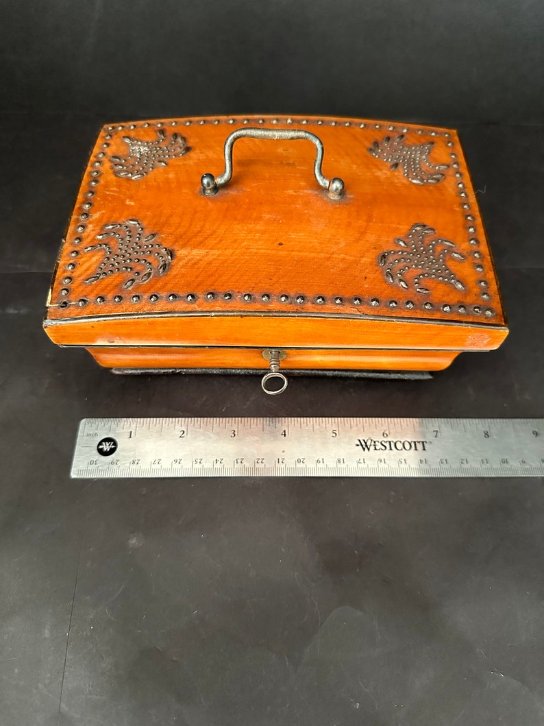 Pu&ograve; includere: Una scatola di legno con un coperchio incernierato, una maniglia in metallo e un buco della serratura in metallo. La scatola &egrave; decorata con borchie in metallo e un disegno di fiori stilizzati. La scatola &egrave; marrone e ha una finitura in legno naturale.
