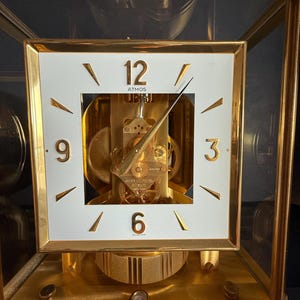 May include: A gold-toned Atmos clock with a white square face. The clock face features black numerals at 3, 6, 9, and 12 o'clock, with gold hands. The clock mechanism is visible through the face. The clock is encased in a glass box.