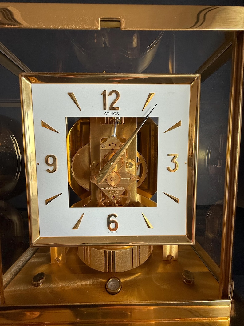 May include: A gold-toned Atmos clock with a white square face. The clock face features black numerals at 3, 6, 9, and 12 o'clock, with gold hands. The clock mechanism is visible through the face. The clock is encased in a glass box.