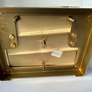 Puede incluir: La parte inferior de una caja de metal de color lat&oacute;n con cuatro tornillos y dos varillas met&aacute;licas. La caja tiene una etiqueta que dice "The Camera Co. of New York" y "Made in U.S.A."