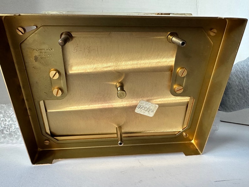 Puede incluir: La parte inferior de una caja de metal de color lat&oacute;n con cuatro tornillos y dos varillas met&aacute;licas. La caja tiene una etiqueta que dice "The Camera Co. of New York" y "Made in U.S.A."