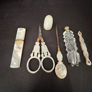 Pu&ograve; includere: Un set di strumenti da cucito vintage in madreperla e vetro. Il set comprende un ditale, delle forbici, un infila-ago, un astuccio per aghi e una piccola bottiglia.