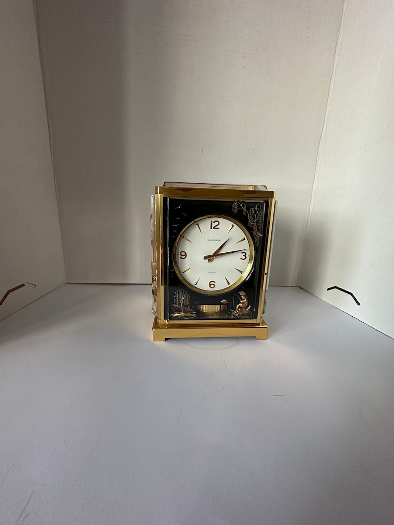 Puede incluir: Un reloj dorado con una esfera negra y agujas blancas. El reloj tiene una parte delantera de vidrio y un dise&ntilde;o decorativo en la parte posterior.