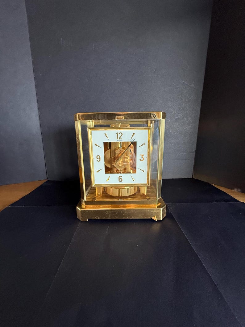 May include: A gold-toned rectangular clock with a white face and black numerals. The clock's face displays the numbers 12, 3, 6, and 9. The clock is encased in glass and has a visible pendulum.