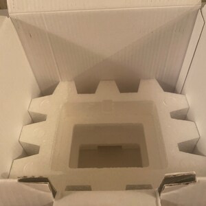 Puede incluir: Una caja de cart&oacute;n blanca abierta con un inserto de poliestireno. El inserto tiene un recorte rectangular en el centro.