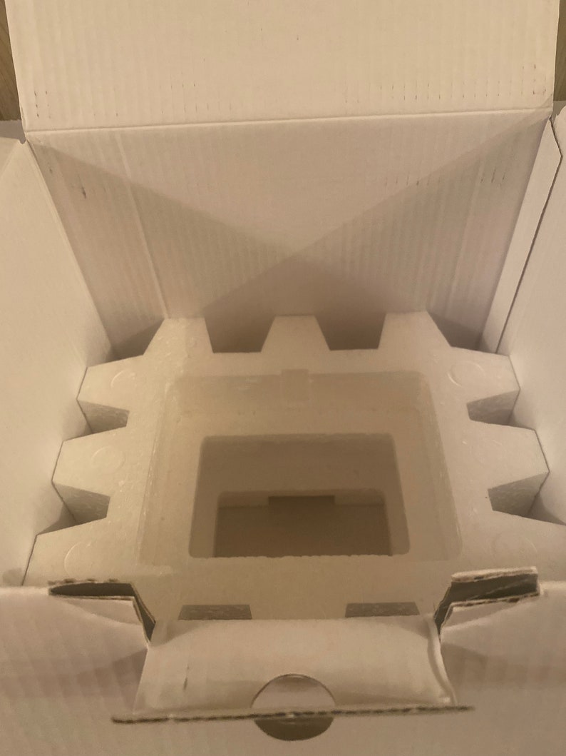 Puede incluir: Una caja de cart&oacute;n blanca abierta con un inserto de poliestireno. El inserto tiene un recorte rectangular en el centro.