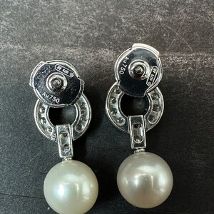 May include: A pair of silver earrings with a pearl drop. The earrings have a circular design with small diamonds around the edge. The back of the earrings are marked with 'AU 750' and '925'.