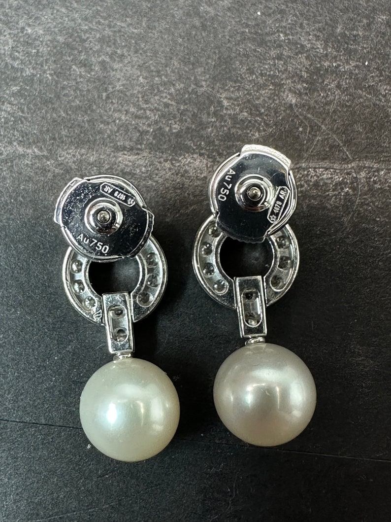 May include: A pair of silver earrings with a pearl drop. The earrings have a circular design with small diamonds around the edge. The back of the earrings are marked with 'AU 750' and '925'.
