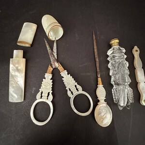 Pu&ograve; includere: Un set di strumenti da cucito vintage, tra cui un paio di forbici con manico di perla, un ditale, un astuccio per aghi, una piccola bottiglia e un piccolo strumento con manico di perla.