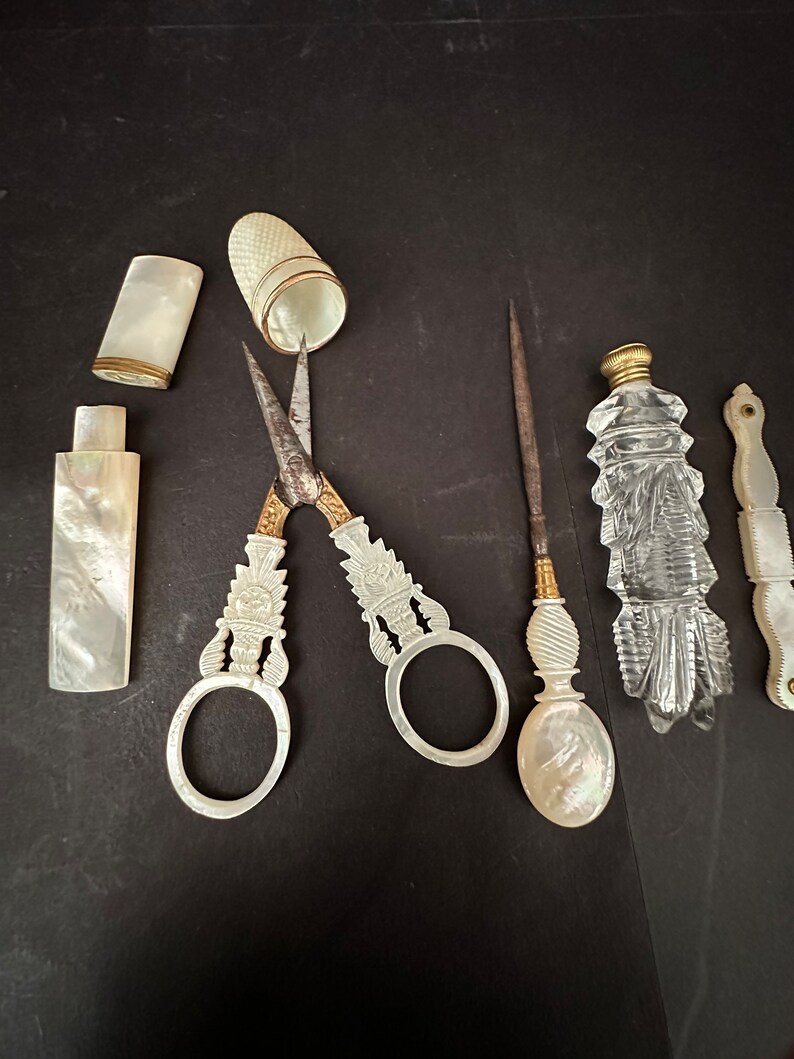 Pu&ograve; includere: Un set di strumenti da cucito vintage, tra cui un paio di forbici con manico di perla, un ditale, un astuccio per aghi, una piccola bottiglia e un piccolo strumento con manico di perla.