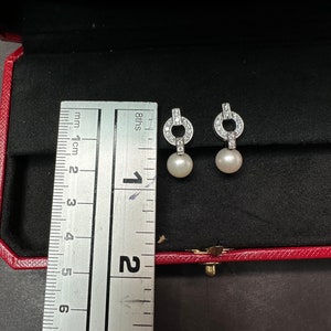 May include: A pair of silver diamond and pearl earrings. The earrings are in a red box with a ruler for size reference. The ruler shows that the earrings are about 1.5 cm long.