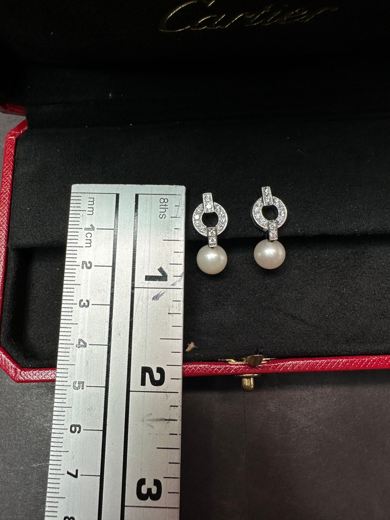 May include: A pair of silver diamond and pearl earrings. The earrings are in a red box with a ruler for size reference. The ruler shows that the earrings are about 1.5 cm long.